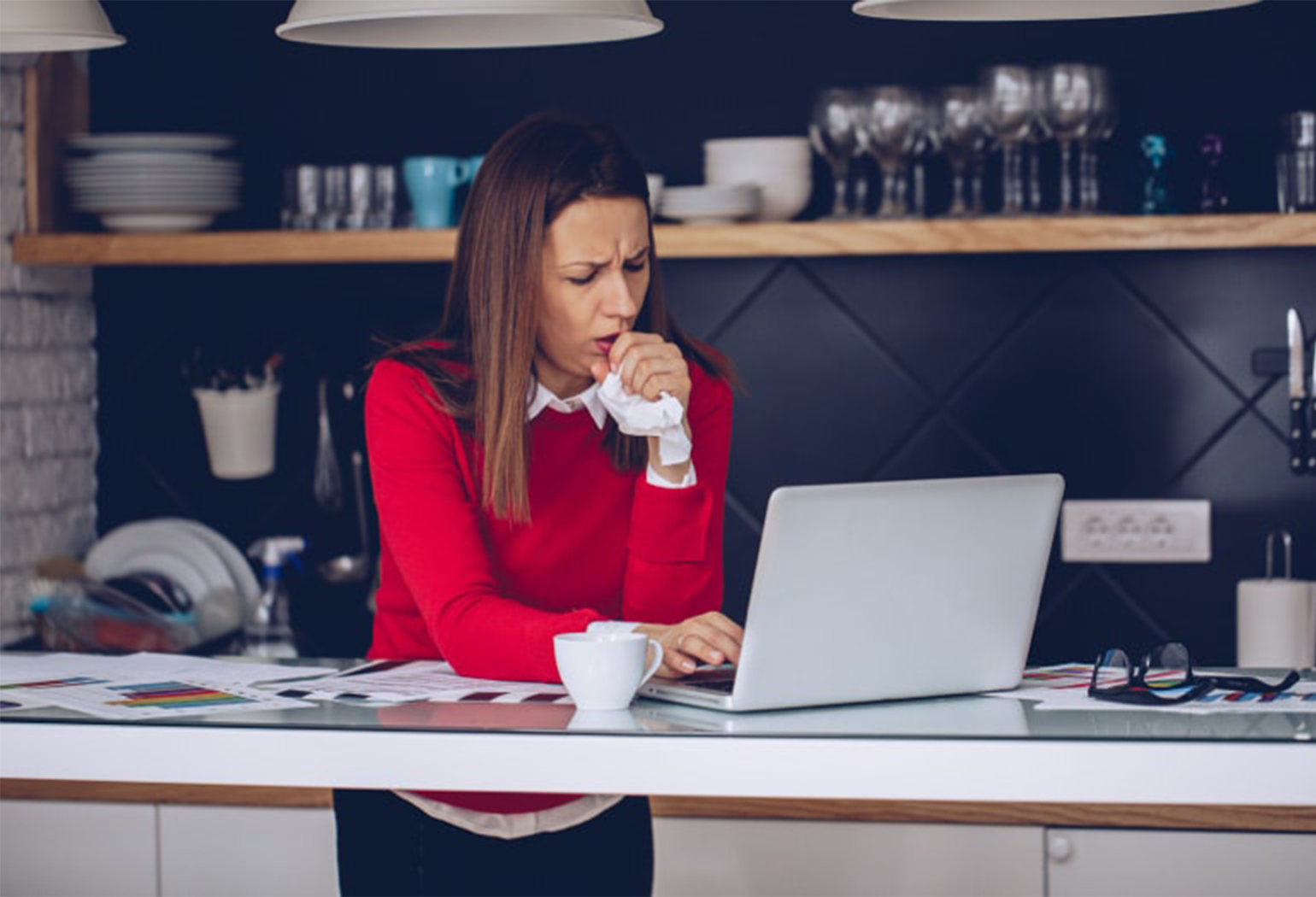 Što uzrokuje kašalj? Žena stoji u kuhinji i radi na prijenosnom računalu dok kašlje u maramicu.